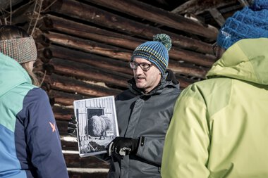 Ein Mann erklärt eine historische Fotografie vor einer Gruppe bei einer Kulturführung im Winter. | © Kleinwalsertal Tourismus eGen • Fotograf: Dominik Berchtold