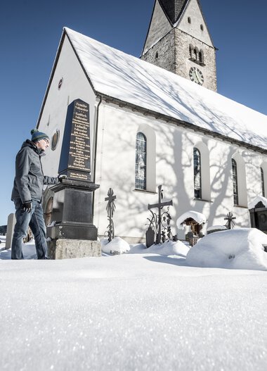 Drei Personen stehen vor einer alten Kirche im verschneiten Winter mit klar blauem Himmel. | © Kleinwalsertal Tourismus eGen • Fotograf: Dominik Berchtold