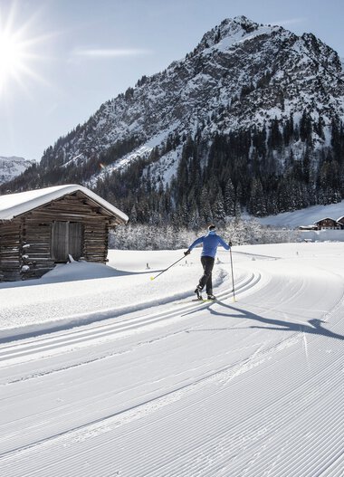 Skifahrer auf einer Langlaufloipe in einer verschneiten Berglandschaft bei Sonnenschein, mit einer kleinen Holzhütte im Vordergrund. | © Kleinwalsertal Tourismus eGen • Fotograf: Dominik Berchtold