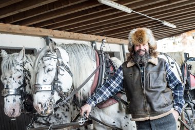 Mann mit Pelzmütze und Lederjacke steht vor zwei weißen Pferden in einer Pferdestallhalle, hält die Leine eines Pferdes und lächelt. | © Kleinwalsertal Tourismus eGen • Fotograf: Dominik Berchtold