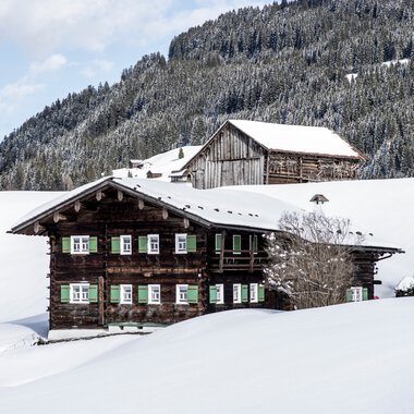 Traditionelles Holzhaus in schneebedeckter Winterlandschaft mit Bergen im Hintergrund. | © Kleinwalsertal Tourismus eGen • Fotograf: Dominik Berchtold