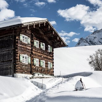 Ein verschneites Holzhaus in den Bergen mit einem kleinen Baum daneben, umgeben von Schnee, unter einem bewölkten Himmel. | © Kleinwalsertal Tourismus eGen • Fotograf: Dominik Berchtold