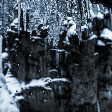 Eiszapfen hängen von einem Dach über einem winterlichen Fluss in der Breitachklamm, mit Schneeschauern im Hintergrund. | © Kleinwalsertal Tourismus eGen • Fotograf: Dominik Berchtold