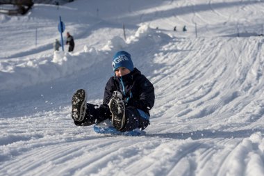 Ein Kind sitzt lachend auf einem Rodel im Schnee, beim Wintershooting auf Mittelberg, umgeben von verschneiten Bergen. | © Kleinwalsertal Tourismus eGen • Fotograf: Dominik Berchtold