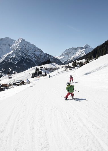 Familie beim Skifahren auf einer verschneiten Piste in den Bergen, umgeben von Winterlandschaft und Talhütten. | © Kleinwalsertal Tourismus eGen • Fotograf: Dominik Berchtold