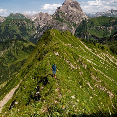A hiker walkers along a lush green mountain ridge with rugged peaks in the background under a partly cloudy sky. | © Kleinwalsertal Tourismus eGen • Fotograf: Erika Dürr