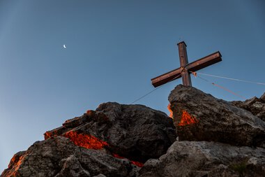 Ein Holzkreuz auf einem Felsen, im Hintergrund der Abendhimmel mit Mondsichel und tiefstehender Sonne, Panorama im Kleinwalsertal. | © Kleinwalsertal Tourismus eGen • Fotograf: Erika Dürr