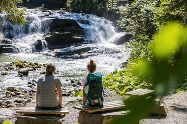 Zwei Frauen sitzen am Flussufer vor einem Wasserfall in einem Wald, umgeben von grünen Büschen und Bäumen. | © Kleinwalsertal Tourismus eGen • Fotograf: Frank Drechsel