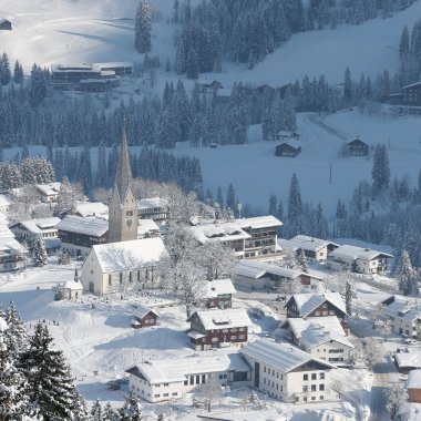 Winteransicht eines verschneiten Dorfes in den Bergen mit Kirche, umgeben von Tannenwald, bei Schneefall. | © Kleinwalsertal Tourismus eGen • Fotograf: Frank Drechsel