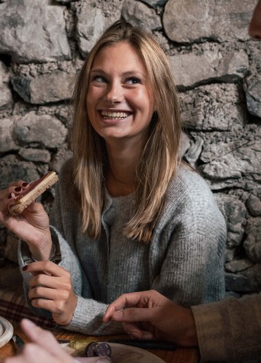Eine junge Frau mit blonden Haaren lächelt in einer gemütlichen, steinernen Umgebung, während sie mit einem Mann spricht und eine kleine Holzfigur hält. | © Kleinwalsertal Tourismus eGen • Fotograf: Hannah Bichay