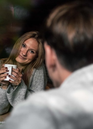 Eine junge Frau hält eine Tasse und lächelt freundlich, während sie einem Mann gegenübersitzt, der nur teilweise sichtbar ist, in einer gemütlichen, privaten Umgebung. | © Kleinwalsertal Tourismus eGen • Fotograf: Hannah Bichay