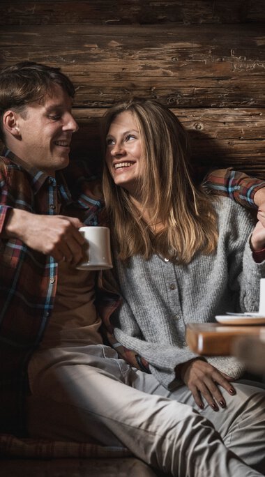 Ein Mann und eine Frau sitzen gemütlich in einer Holzhütte, lachen und trinken Kaffee, umgeben von warmem Holzinterieur. | © Kleinwalsertal Tourismus eGen • Fotograf: Hannah Bichay