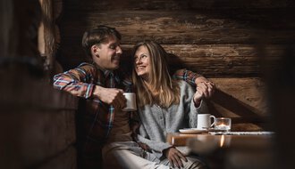 Ein Mann und eine Frau sitzen gemütlich in einer Holzhütte, lachen und trinken Kaffee, umgeben von warmem Holzinterieur. | © Kleinwalsertal Tourismus eGen • Fotograf: Hannah Bichay