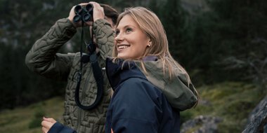 Eine junge Frau mit braunen Haaren und einer blauen Jacke beim Wandern oder Naturerkundung, lächelnd in einer grünen, bewaldeten Umgebung.