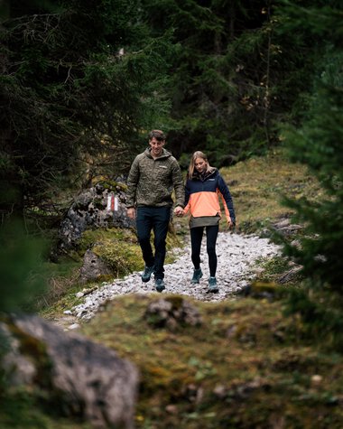 Ein Paar wandert Hand in Hand durch einen bewaldeten Pfad in der Natur bei Tageslicht. | © Kleinwalsertal Tourismus eGen • Fotograf: Hannah Bichay