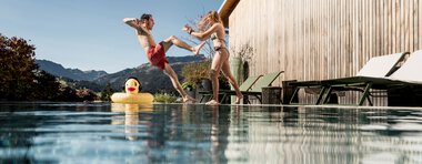 Zwei Kinder springen fröhlich in einen Pool, mit einer Schwimmring-Enten im Wasser, im Hintergrund ein Holzhaus und Berglandschaft bei sonnigem Wetter. | © Kleinwalsertal Tourismus eGen • Fotograf: Hannah Bichay
