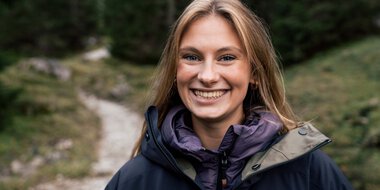 Young woman with long blonde hair, smiling, wearing an outdoor jacket, standing in a forested area with a stream in the background during a photoshoot in nature. | © Kleinwalsertal Tourismus eGen • Photographer: Hannah Bichay