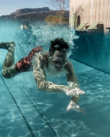 Ein Mann taucht in einem Wasserbecken mit schnellen Bewegungen und Wasserspritzern, während im Hintergrund Berge und ein Pavillon sichtbar sind. | © Kleinwalsertal Tourismus eGen • Fotograf: Hannah Bichay