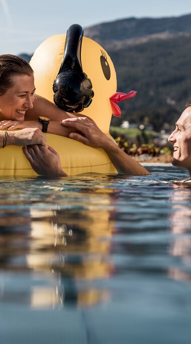 Paare schwimmen im See mit einem großen gelben Gummiente-Schwimmring, lachen und genießen das schöne Wetter vor Bergen im Hintergrund. | © Kleinwalsertal Tourismus eGen • Fotograf: Hannah Bichay