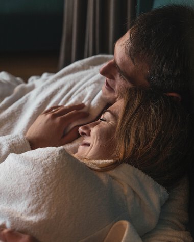 Ein glückliches Paar liegt im Bett, der Mann umarmt die Frau, beide lachen und wirken entspannt und verliebt. | © Kleinwalsertal Tourismus eGen • Fotograf: Hannah Bichay