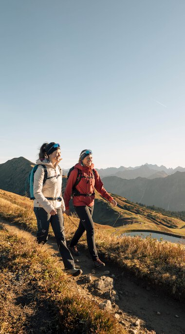 Zwei Wanderer auf einem schmalen Pfad in den Bergen mit einem See im Hintergrund, sonniges Wetter, Panorama der Berglandschaft. | © Kleinwalsertal Tourismus eGen • Fotograf: Hannah Bichay