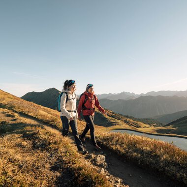 Zwei Wanderer auf einem schmalen Pfad in den Bergen mit einem See im Hintergrund, sonniges Wetter, Panorama der Berglandschaft. | © Kleinwalsertal Tourismus eGen • Fotograf: Hannah Bichay