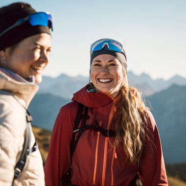 Zwei lachende Frauen beim Wandern in den Bergen, gekleidet in Outdoor-Bekleidung und Sonnenbrillen.