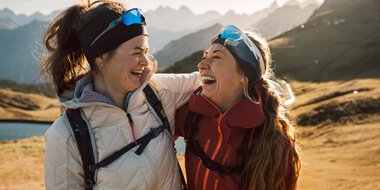 Zwei lachende Frauen beim Wandern in den Bergen, umgeben von einer malerischen Landschaft mit Bergen im Hintergrund.