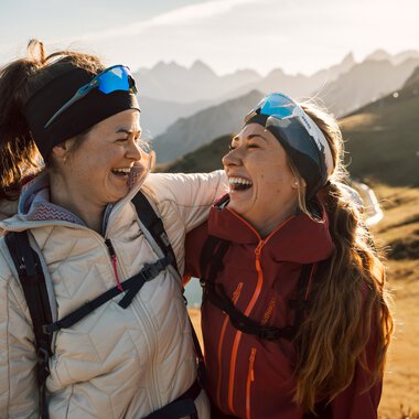 Zwei lachende Frauen beim Wandern in den Bergen, umgeben von einer malerischen Landschaft mit Bergen im Hintergrund.