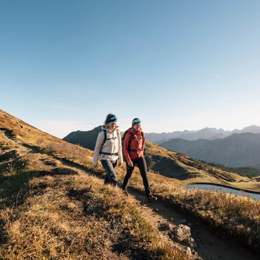 Zwei Wanderer auf einem Pfad in den Bergen bei Sonnenuntergang, mit einer Berglandschaft und einem See im Hintergrund. | © Kleinwalsertal Tourismus eGen • Fotograf: Hannah Bichay