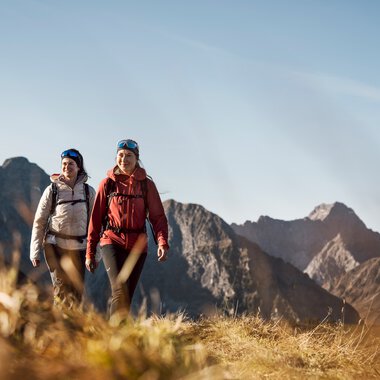 Zwei Frauen wandern in den Bergen bei sonnigem Wetter, mit Rucksäcken und sportlicher Kleidung.