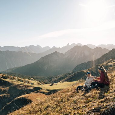 Zwei Personen sitzen entspannt auf einer Bergwiese mit Blick auf eine beeindruckende Bergkette bei Sonnenuntergang.