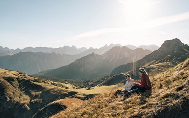 Two people sitting relaxed on a mountain meadow with a view of a stunning mountain range at sunset.
