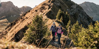 Zwei wandernde Frauen auf einem schmalen Bergpfad mit einer beeindruckenden Bergkulisse im Hintergrund, umgeben von Tannenbäumen.