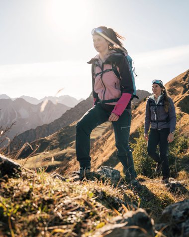 Zwei Frauen wandern auf einem Bergpfad in den Bergen bei Sonnenlicht, umgeben von Natur und felsigen Landschaften.