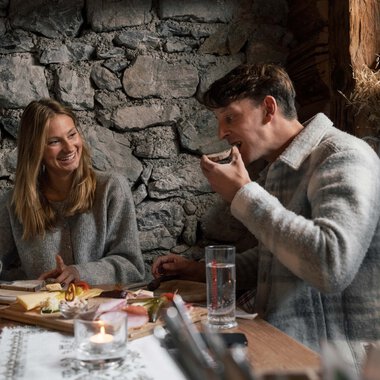 Ein Mann und eine Frau sitzen in einem gemütlichen Raum mit Steinausstattung am Tisch, lachen und genießen ein gemeinsames Gespräch bei Kerzenschein. | © Kleinwalsertal Tourismus eGen • Fotograf: Hannah Bichay