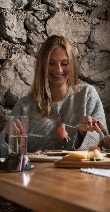 Zwei Menschen sitzen fröhlich beim Essen in einer gemütlichen Holz- und Steingasthütte, lachen und unterhalten sich. | © Kleinwalsertal Tourismus eGen • Fotograf: Hannah Bichay