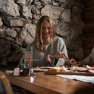 Two people are happily dining in a cozy wooden and stone cabin, laughing and engaging in conversation. | © Kleinwalsertal Tourismus eGen • Photographer: Hannah Bichay