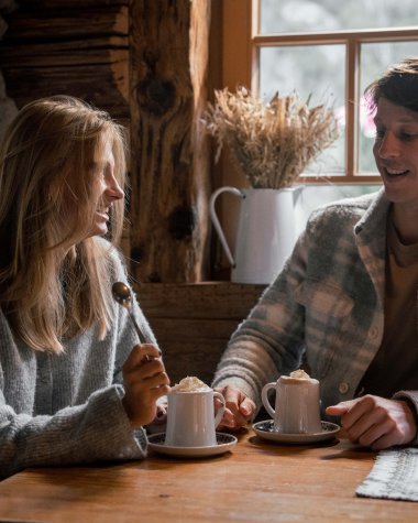 Zwei junge Menschen sitzen sich in einem gemütlichen Café gegenüber, lachen und genießen eine Tasse Kaffee bei Kerzenlicht in einer rustikalen Holz- und Steinumgebung. | © Kleinwalsertal Tourismus eGen • Fotograf: Hannah Bichay