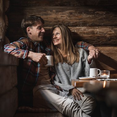 A man and a woman sitting comfortably inside a wooden cabin, laughing and drinking coffee, surrounded by warm wooden interior. | ©  Kleinwalsertal Tourismus eGen • Photographer: Hannah Bichay
