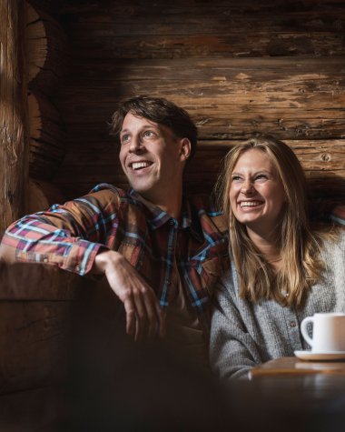 Two happy people sitting inside a cozy wooden cabin, laughing and enjoying a pleasant moment at a table with cups. | © Kleinwalsertal Tourismus eGen • Photographer: Hannah Bichay