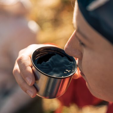Close-up of a person in nature holding and drinking from a cup with hot beverage, in outdoor sunlight. | © Kleinwalsertal Tourismus eGen • Photographer: Hannah Bichay