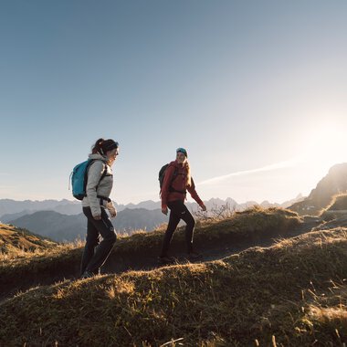 Three hikers with backpacks walking on a narrow trail in a mountain landscape at sunset. | ©  Kleinwalsertal Tourismus eGen • Photographer: Hannah Bichay