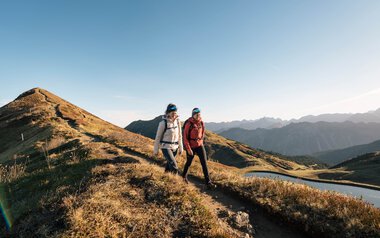 Two hikers walking on a trail in the mountains at sunset, with a mountain landscape and a lake in the background. | © Kleinwalsertal Tourismus eGen • Photographer: Hannah Bichay