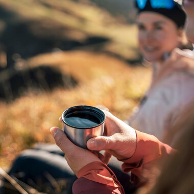 Two people hiking outdoors, one is holding a coffee cup, with a scenic landscape in the background during daylight. | © Kleinwalsertal Tourismus eGen • Photographer: Hannah Bichay