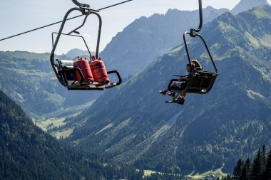 Zwei Personen sitzen in einer Sommer-Skiliftanlage in den Bergen, mit Blick auf eine grüne Almregion und die umliegenden Berge. | © Kleinwalsertal Tourismus eGen • Fotograf: Oliver Farys