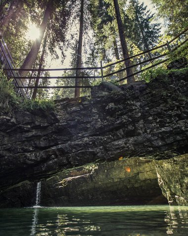 Naturbrücke in einer Höhle mit Wasser, Sonnenlicht durch die Baumkronen, Holzbrücke im Hintergrund. | © Kleinwalsertal Tourismus eGen • Fotograf: Oliver Farys