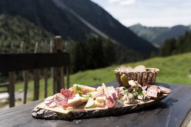 Käse-, Wurst- und Gemüseplatte auf einem Holztisch mit Bergpanorama im Hintergrund, ideal für eine Bergwanderung | © Kleinwalsertal Tourismus eGen • Fotograf: Oliver Farys