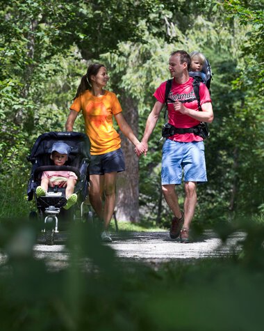 Familie beim Spaziergang im Wald, Mutter, Vater und Kind im Kinderwagen, umgeben von grünen Bäumen. | © Kleinwalsertal Tourismus eGen • Fotograf: Oliver Farys