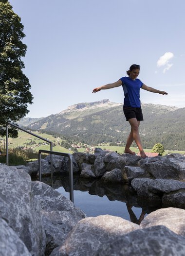 Frau balanciert auf einer natürlichen Steinbrücke in einer Erlebniswelt mit Wasserbecken und Wandermotiv im Hintergrund, umgeben von Bergen und Natur. | © Kleinwalsertal Tourismus eGen • Fotograf: Oliver Farys